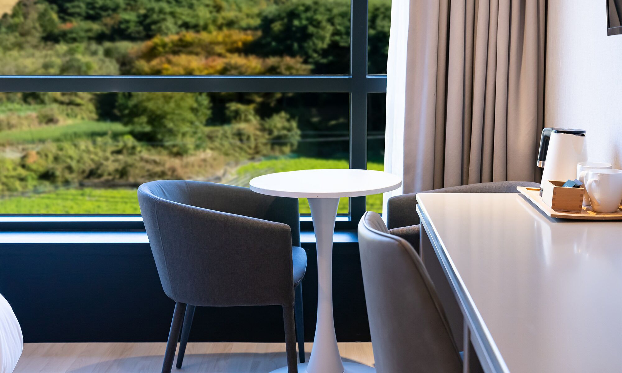 Habitación de hotel con vista a un paisaje verde, una mesa redonda blanca, una silla gris y una cama con ropa de cama blanca.