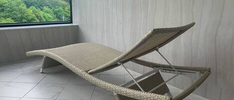Una silla de mimbre con estructura metálica, ubicada en una habitación con piso de baldosas y un ventanal con vistas a un área boscosa.