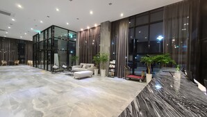 A spacious lobby with a marble floor, a seating area with a sofa and chairs, large windows with curtains, and a reception desk with a plant.