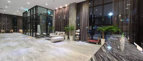 Un vestíbulo amplio con suelo de mármol, zona de estar con sofá y sillones, ventanas grandes con cortinas y un mostrador de recepción con una planta.