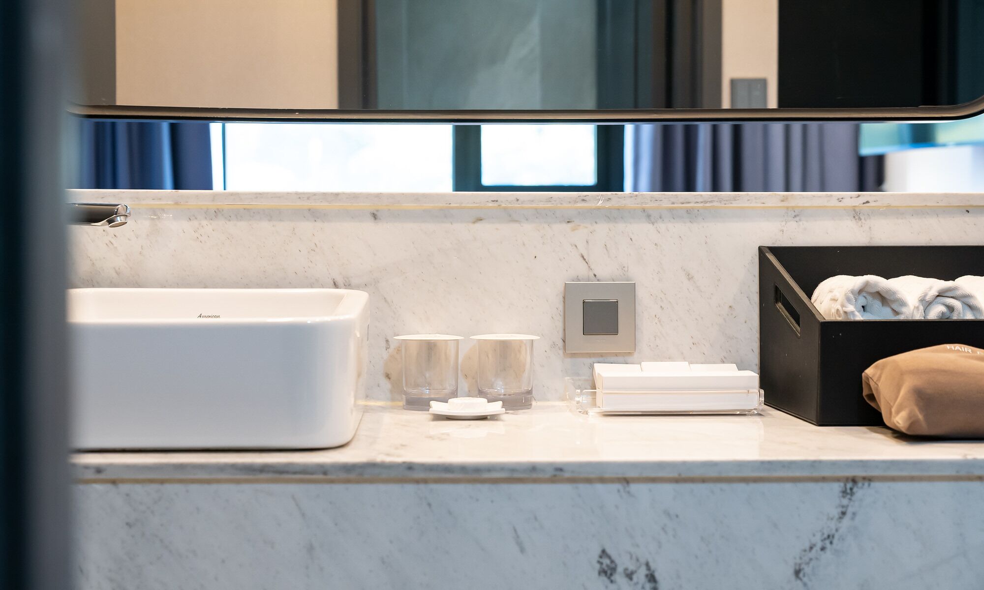 A modern bathroom with a large white sink, two glass tumblers, a towel rack, and a black storage box with towels.