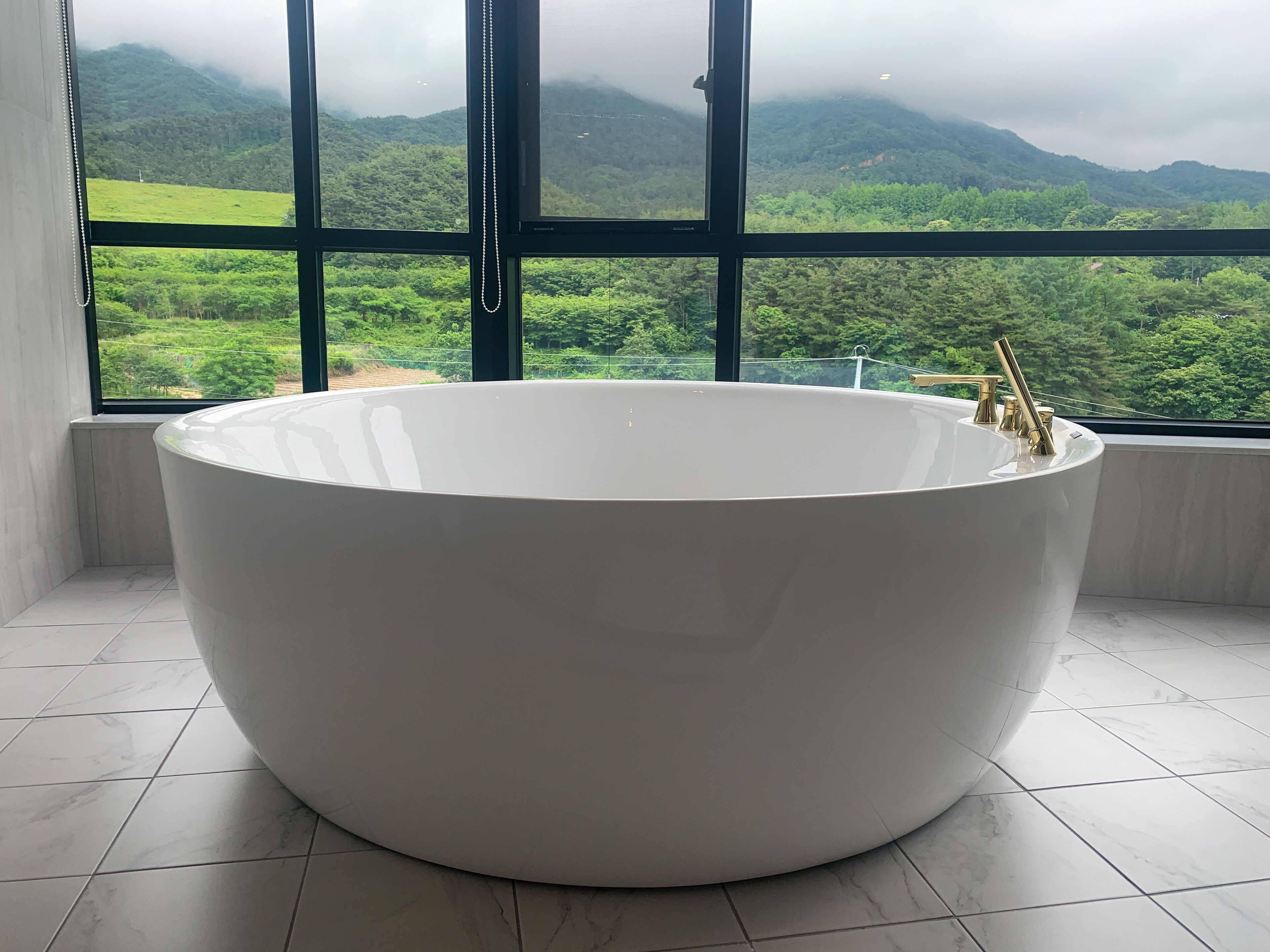 A large, white freestanding bathtub in a bathroom with a view of lush green hills through large windows.