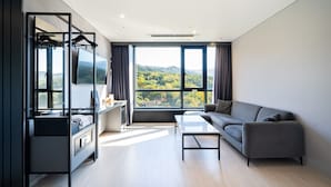 A modern hotel room with a sofa, a coffee table, a TV, and a desk with a computer and a flat-screen TV. Large windows offer a view of a mountainous landscape.