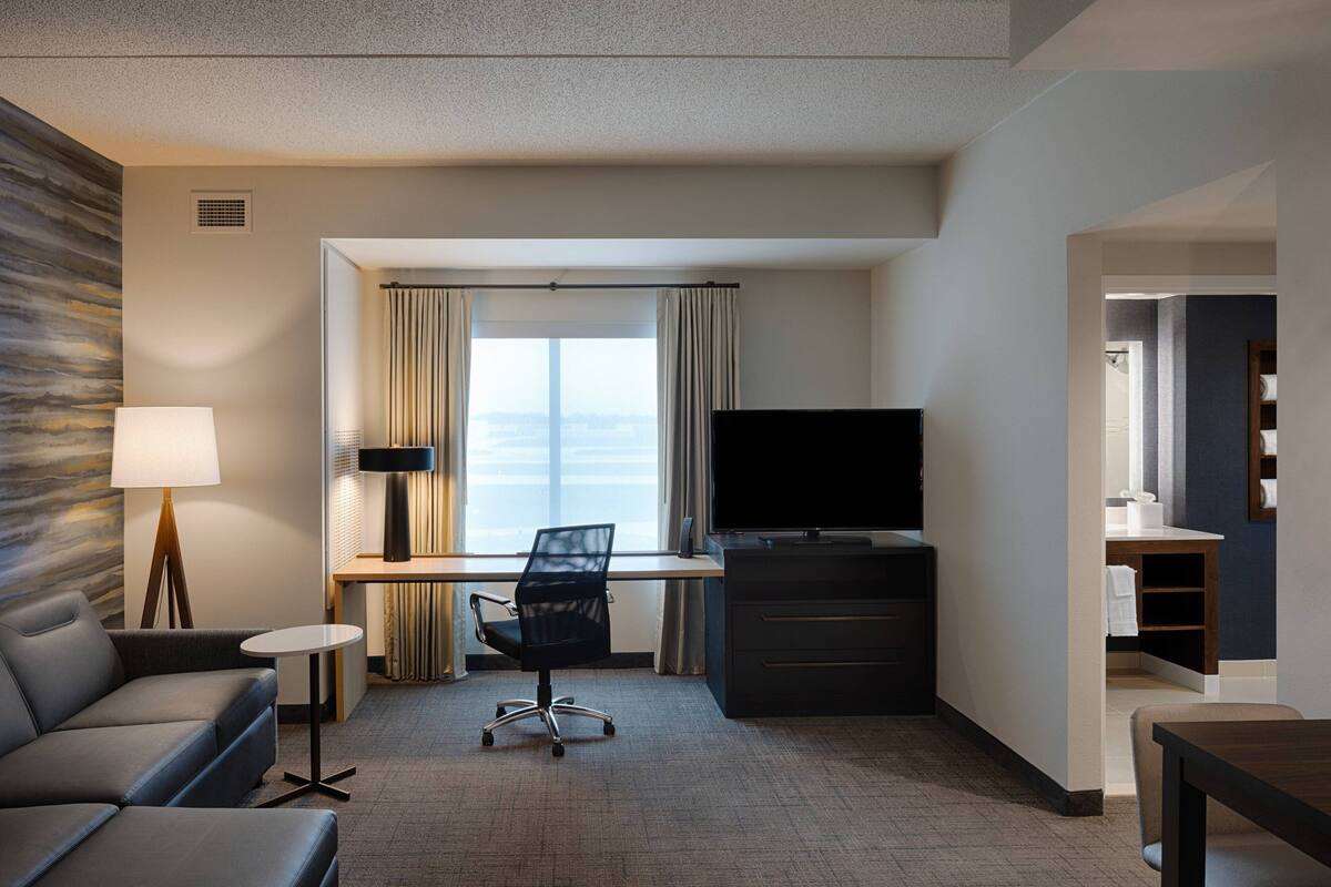 Habitación de hotel con sofá, escritorio, silla, televisor y vista a la ciudad.