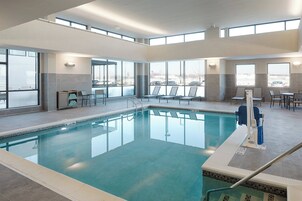 Una piscina interior con paredes de vidrio transparente, piso de baldosas y zona junto a la piscina con sillas y una mesa.