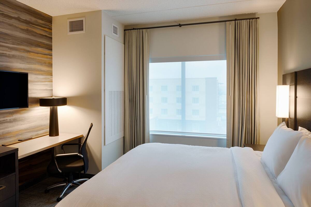 Habitación de hotel con una cama grande, un escritorio con una silla, un televisor y una ventana con cortinas.