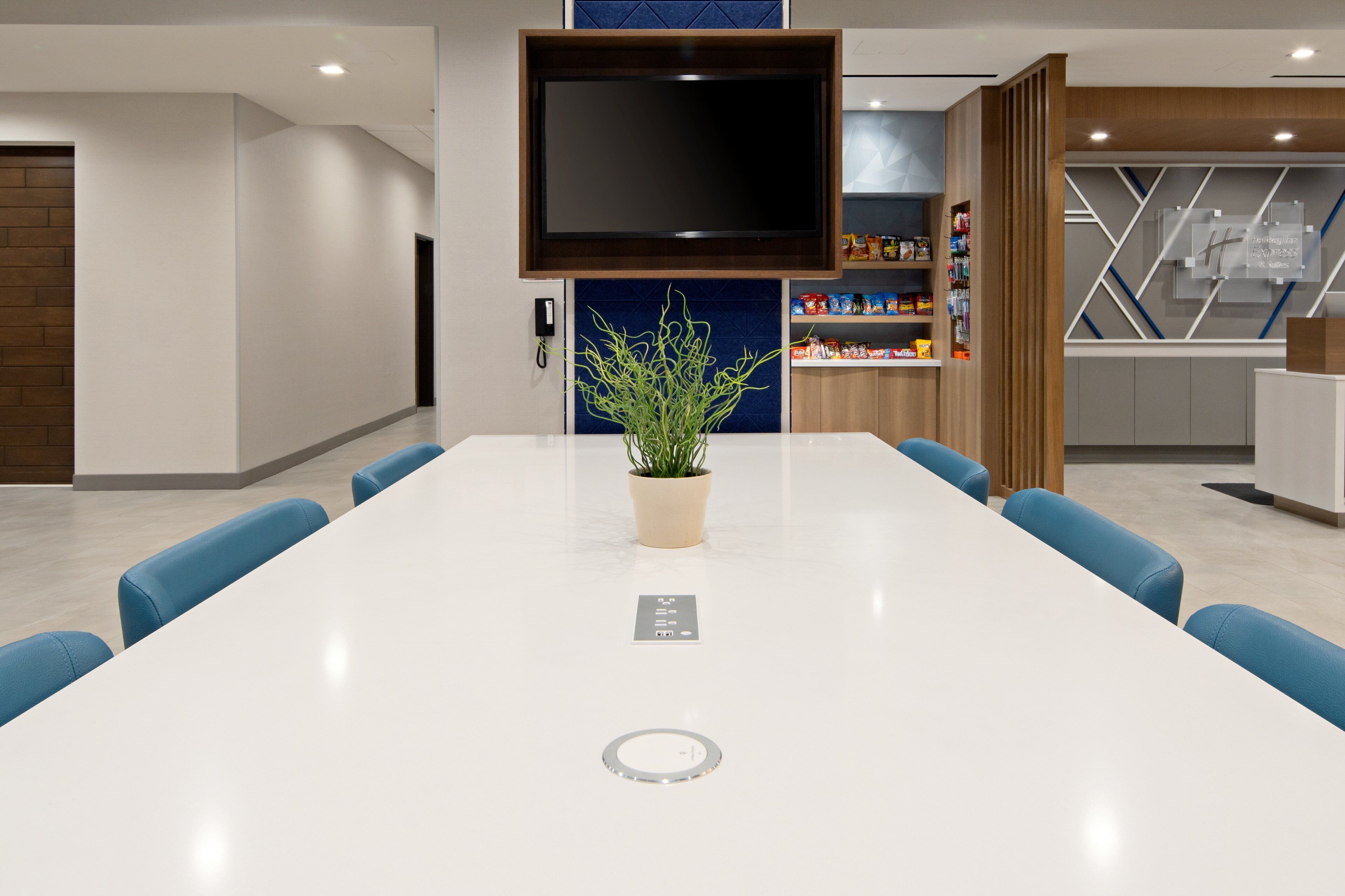 A modern conference room with a white table, blue chairs, a potted plant, a flat-screen TV, and a blue accent wall.