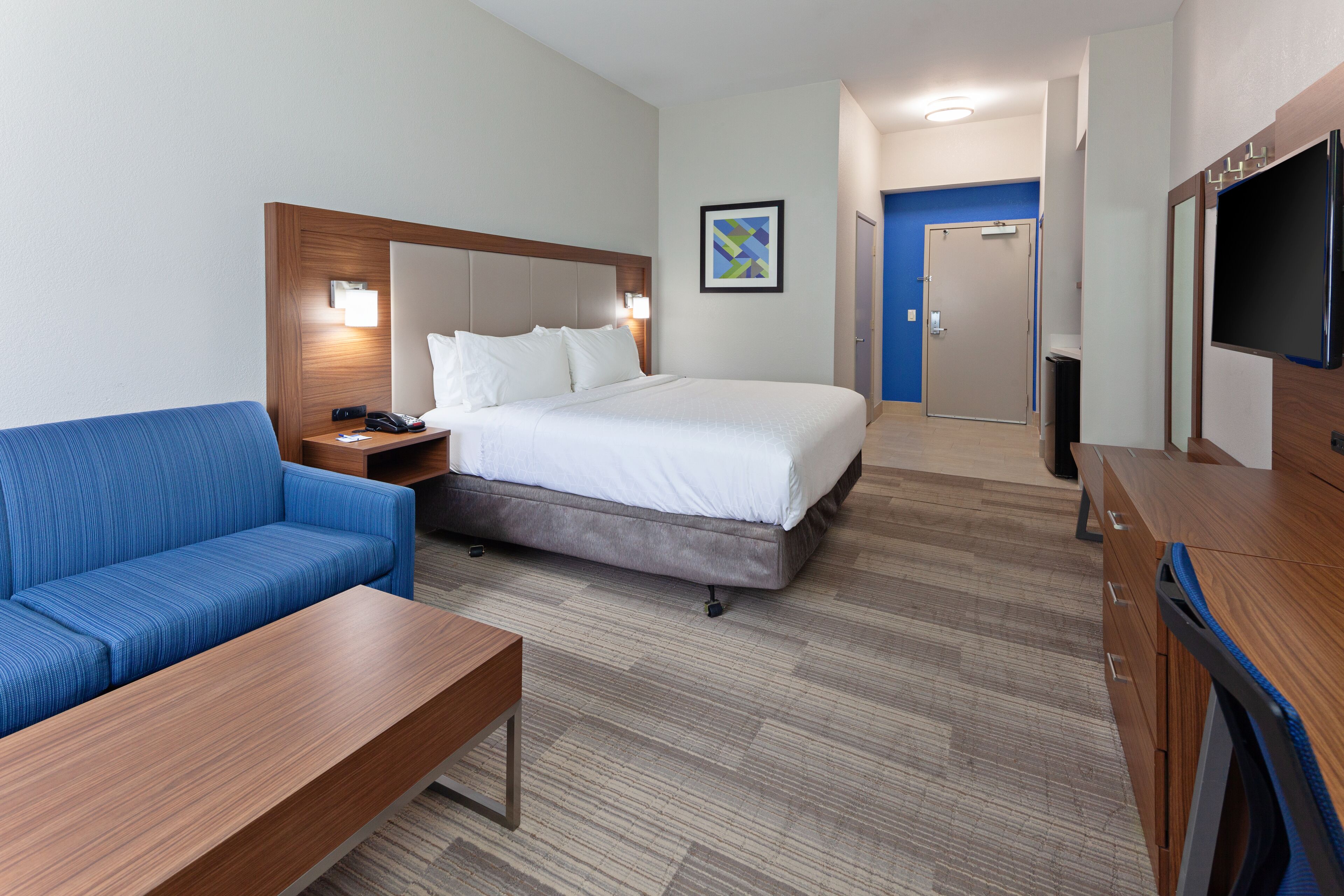 A hotel room with a bed, a blue sofa, a wooden table, a TV, and a door leading to a bathroom.