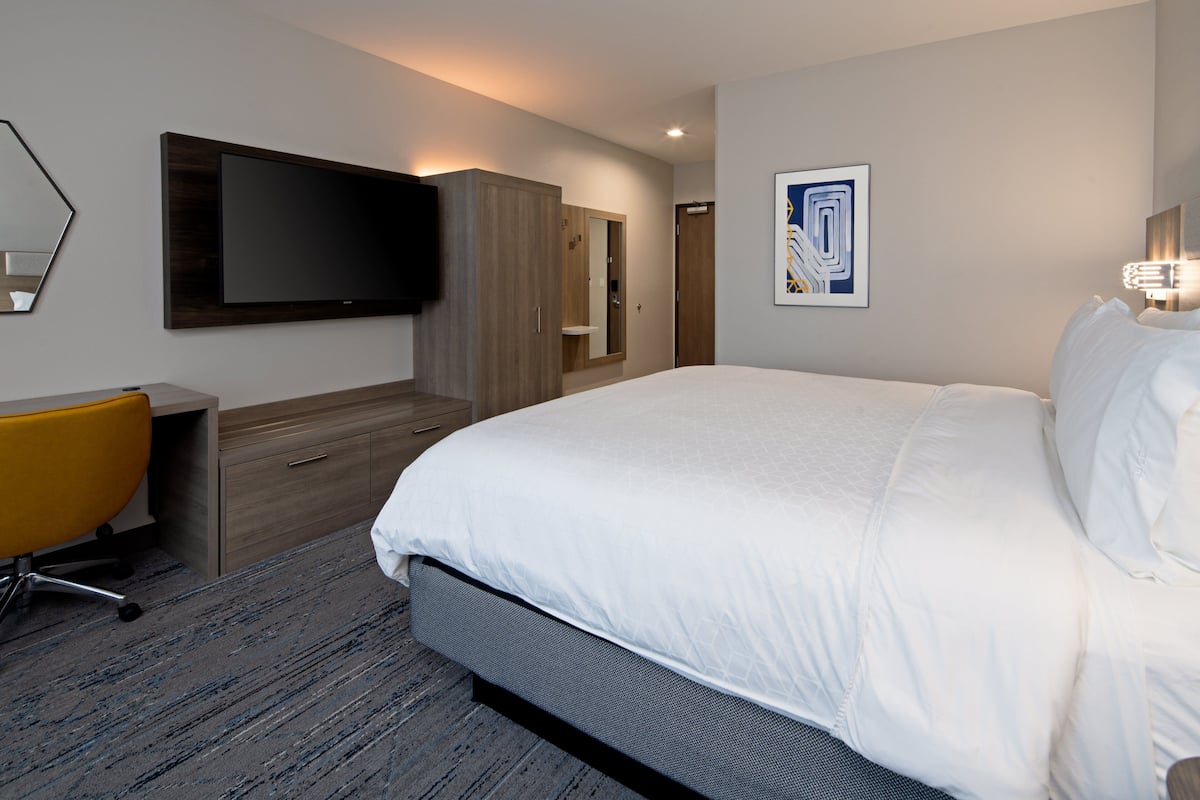 A modern hotel room with a large bed, a flat-screen TV mounted on the wall, a wooden wardrobe, a desk with a chair, and a framed artwork on the wall.
