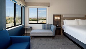Habitación de hotel con una cama grande, un sofá azul, un sillón pequeño, una mesa redonda y vistas a una autopista y montañas a través del ventanal.