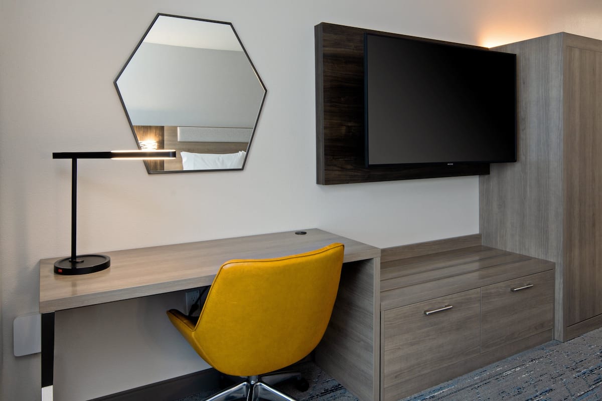 A modern hotel room with a wooden desk, a yellow chair, a flat-screen TV, and a large mirror.