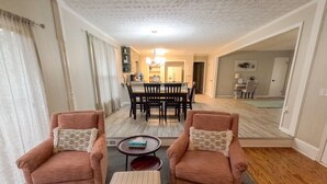 Ein Wohnzimmer mit zwei Sesseln, einem Pouf und einem Essbereich mit Tisch und Stühlen.