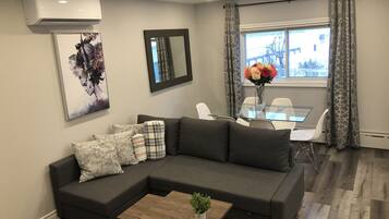 A modern living room with a sectional sofa, a glass coffee table, a dining table with chairs, and a large window with curtains.