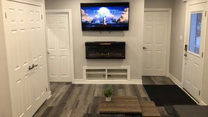 A modern living room with a flat-screen TV mounted above a fireplace, white doors, and a wooden coffee table with a plant. - Stunning Niagara Warm,Cozy and Clean Suite Stay (2) (Welland)