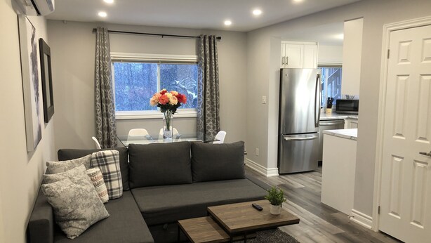 A modern living room with a grey sectional sofa, a small wooden coffee table, and a vase with flowers. A window with curtains is visible, and a kitchen area with a stainless steel refrigerator is in the background. - Stunning Niagara Warm,Cozy and Clean Suite Stay (2) (Welland)