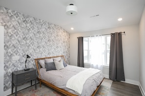 Un dormitorio con pared de estampado geométrico, cama con estructura de madera, mesita de noche con lámpara y ventana con cortinas.