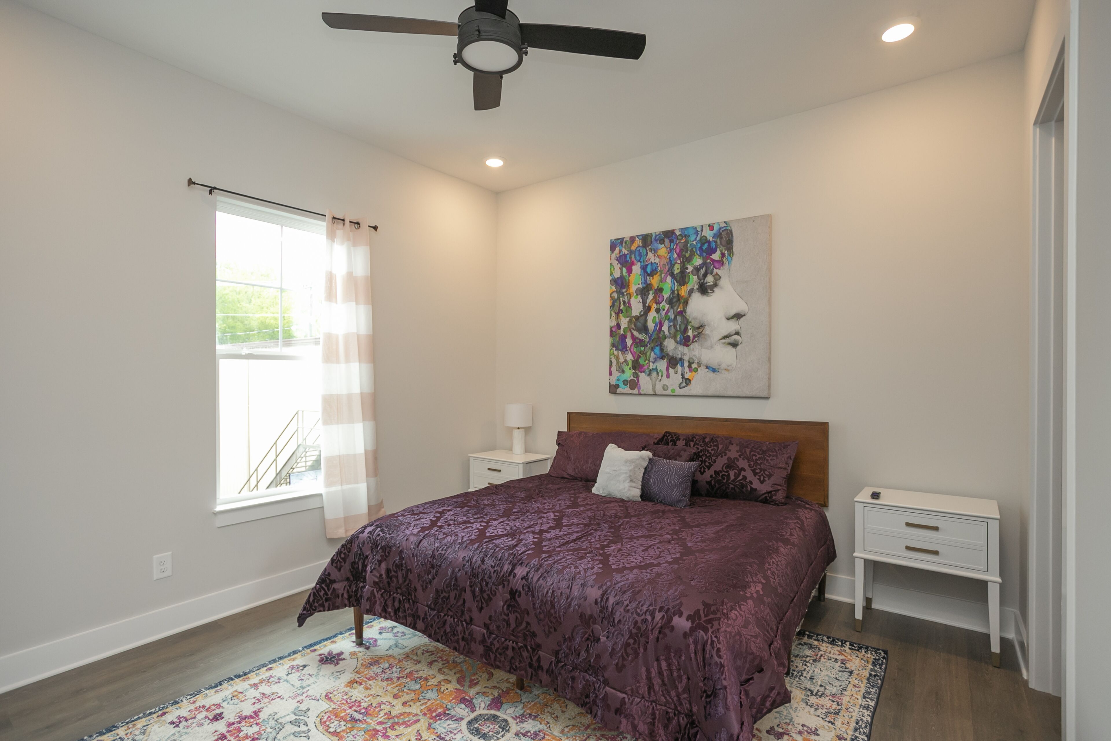 A bedroom with a bed, a ceiling fan, a window with curtains, a nightstand, and a painting on the wall.