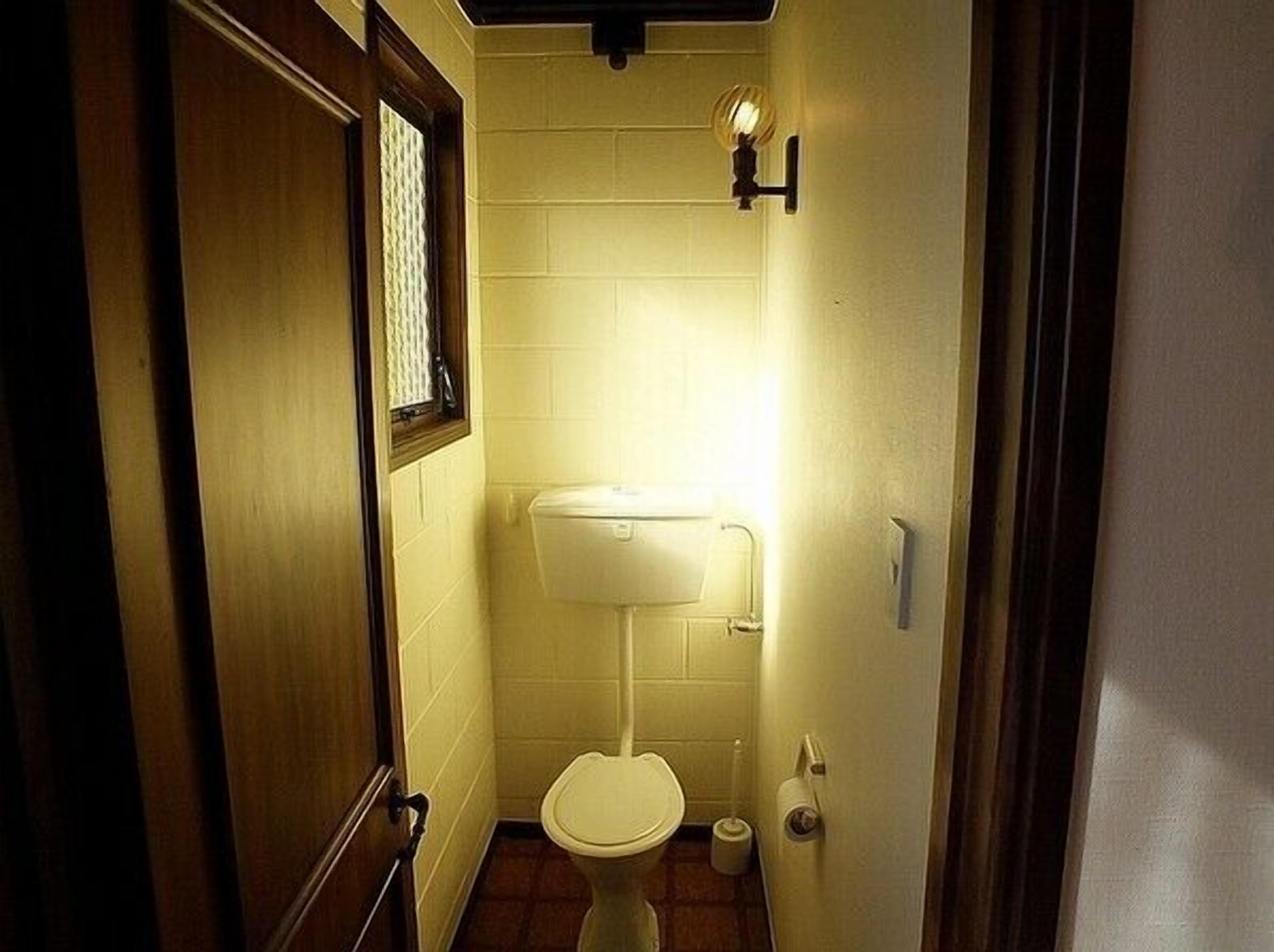Un baño pequeño y estrecho con inodoro, una lámpara empotrada en la pared y una ventana con cortina estampada.