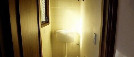 Un baño pequeño y estrecho con inodoro, una lámpara empotrada en la pared y una ventana con cortina estampada.
