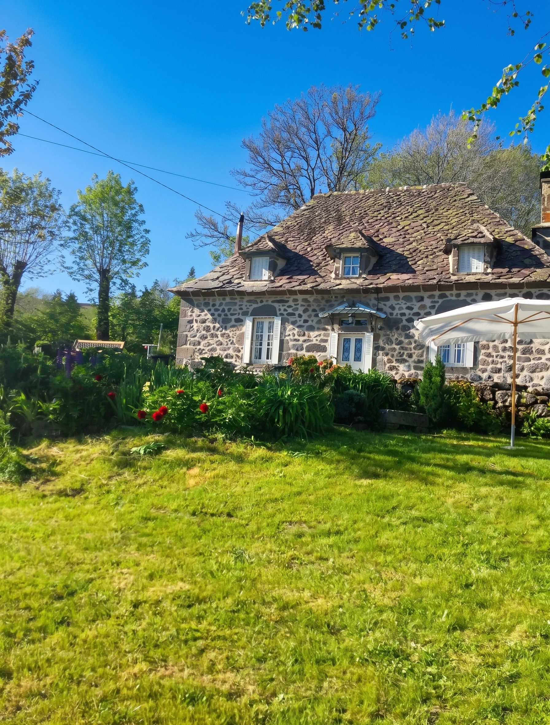 A stone house with a sloped roof, surrounded by a well-maintained garden with various plants and flowers.