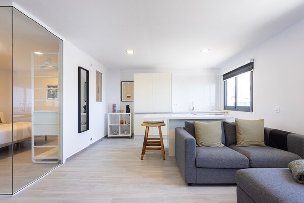 Un apartamento moderno con kitchenette, un sofá gris, un taburete de madera y un separador de vidrio.