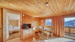 Interior d'una cabana de fusta amb zona de menjador, televisió i vistes a les muntanyes.