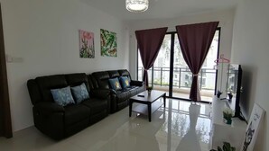 A living room with a black leather sofa, a coffee table, and a TV on a stand. There are two windows with curtains, a chandelier, and artwork on the walls. - Forest City Staview Bay by Wastone (Gelang Patah)