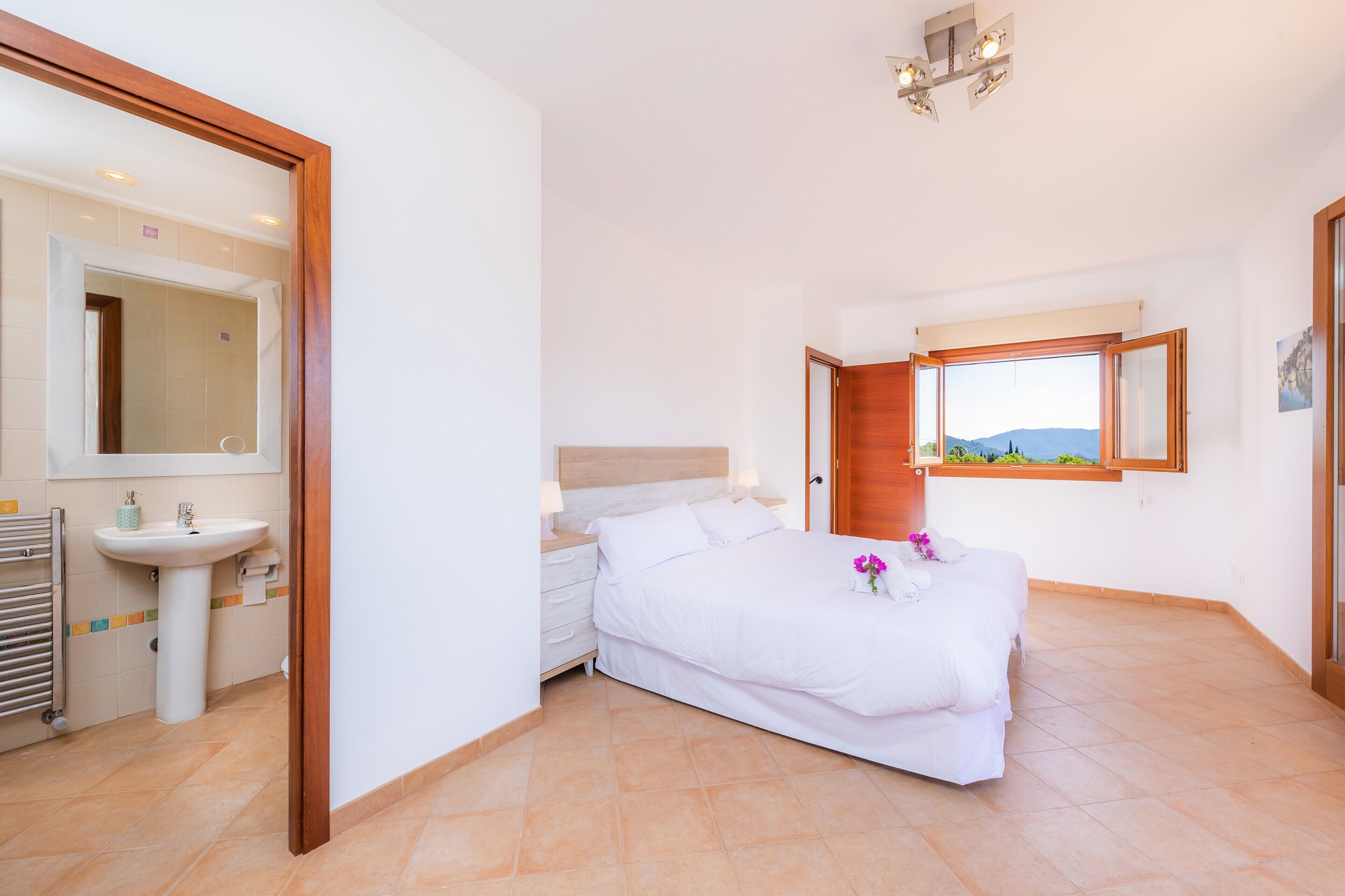 Une chambre à coucher comprenant un lit, une table de chevet, une fenêtre avec vue, une salle de bain avec un lavabo et un miroir, et une porte donnant sur une autre pièce.