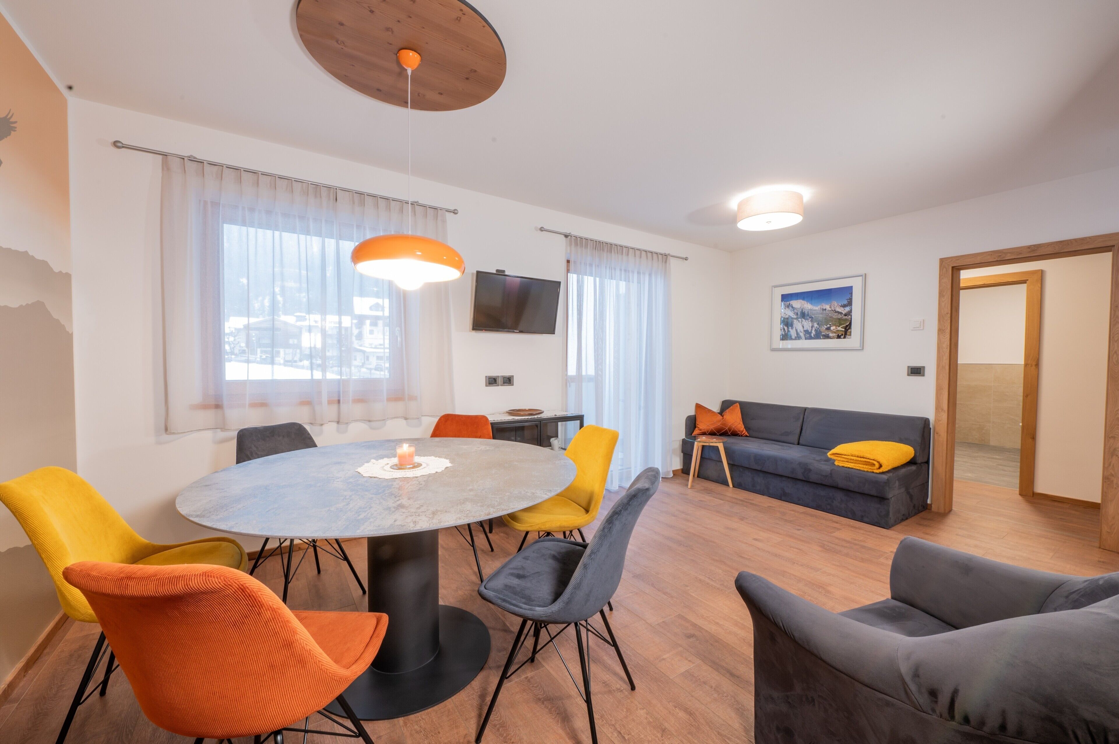 Un salon moderne avec une table de salle à manger ronde, des chaises colorées, un canapé et un téléviseur fixé au mur.