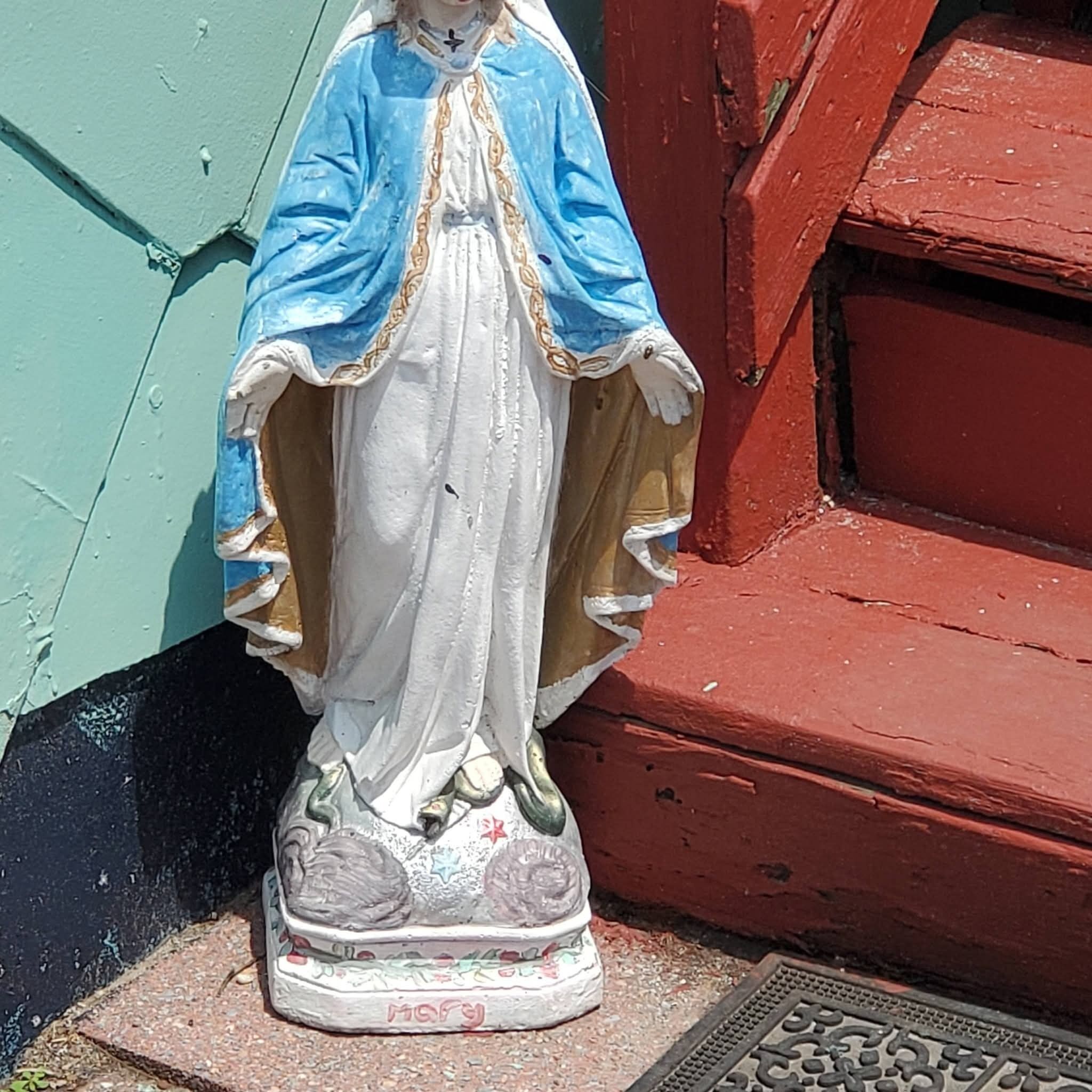 Una statua raffigura una figura con veste bianca e dettagli blu e dorati, posata su un piedistallo decorato con motivi floreali, davanti a un muro verde e rosso.