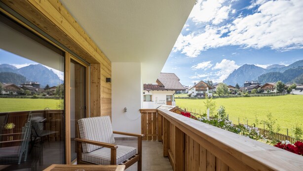 Un balcone in legno con una sedia e un tavolo, vista su un campo verde e case, con montagne sullo sfondo.