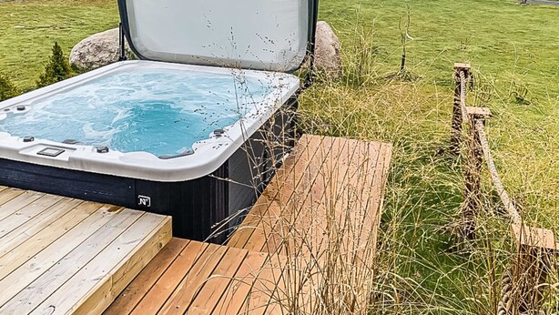 A hot tub with a wooden deck and a covered area, surrounded by a grassy yard and trees.