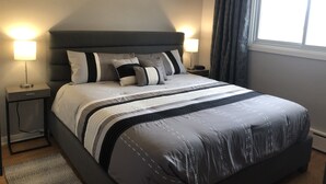 A neatly made bed with a dark headboard, two bedside tables with lamps, and a window with curtains. - Niagara Executive Luxury - HUGE, Warm, Cozy and Clean 3 Bed, 1 Bath SuiteStay(1) (Welland)