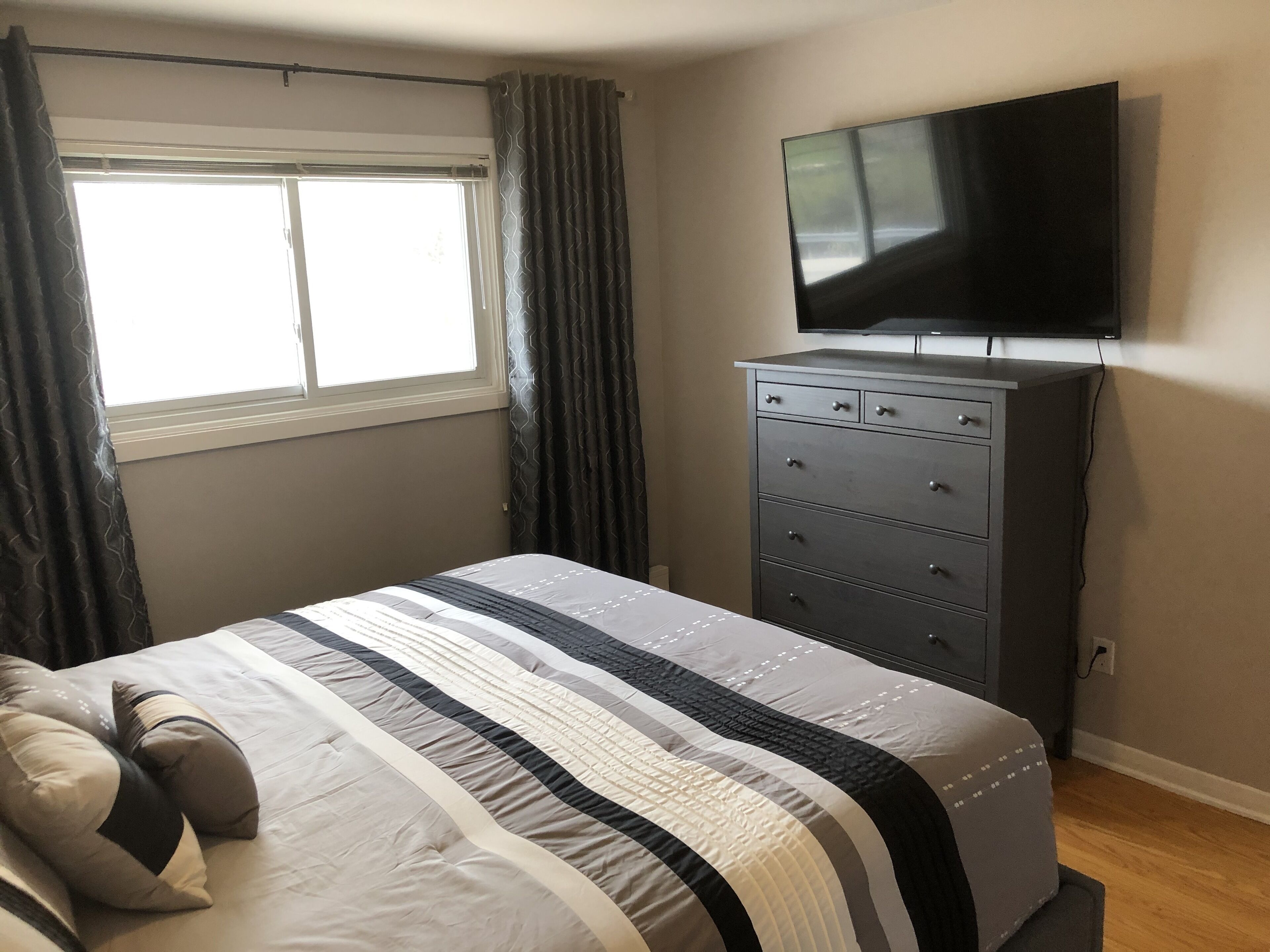 Une chambre à coucher avec un lit, une commode, un téléviseur fixé au mur et une fenêtre avec des rideaux.