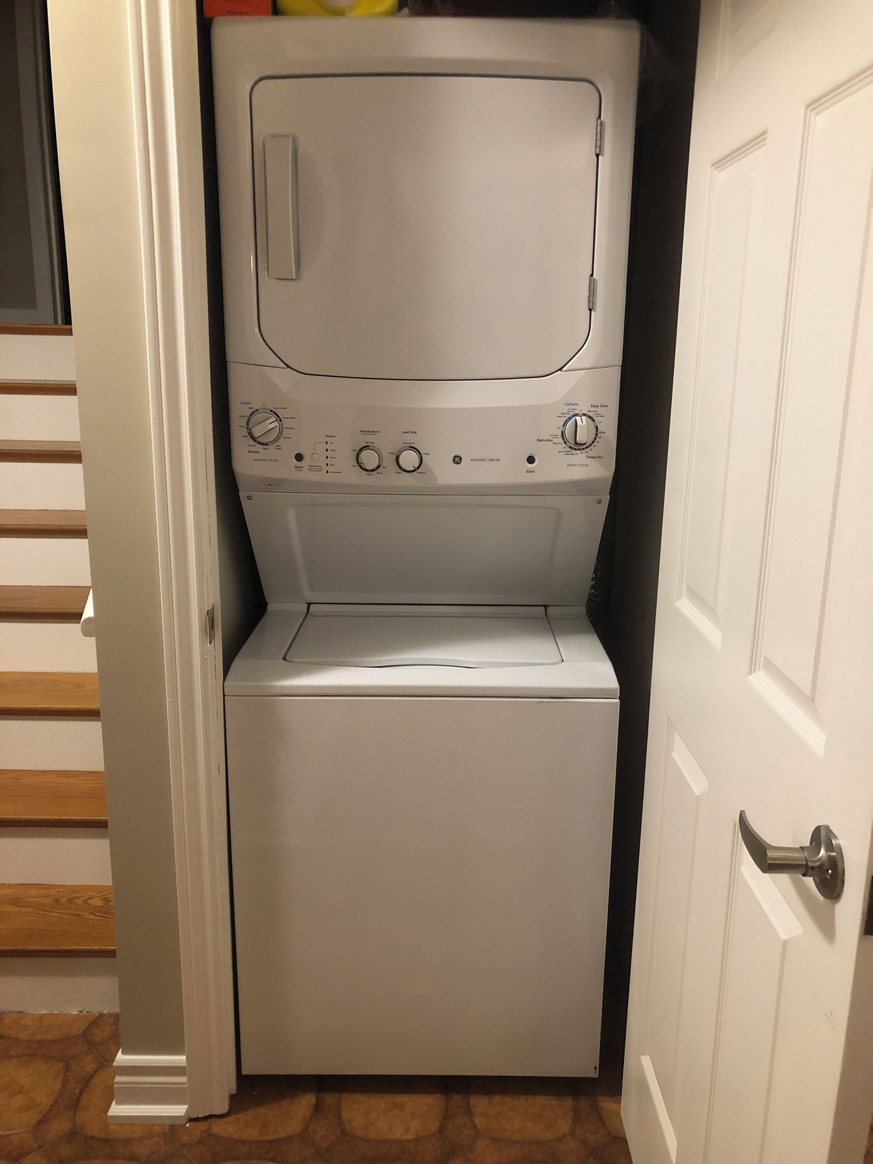 A compact, white, front-loading washing machine in a small laundry room with a wooden floor and a white door.