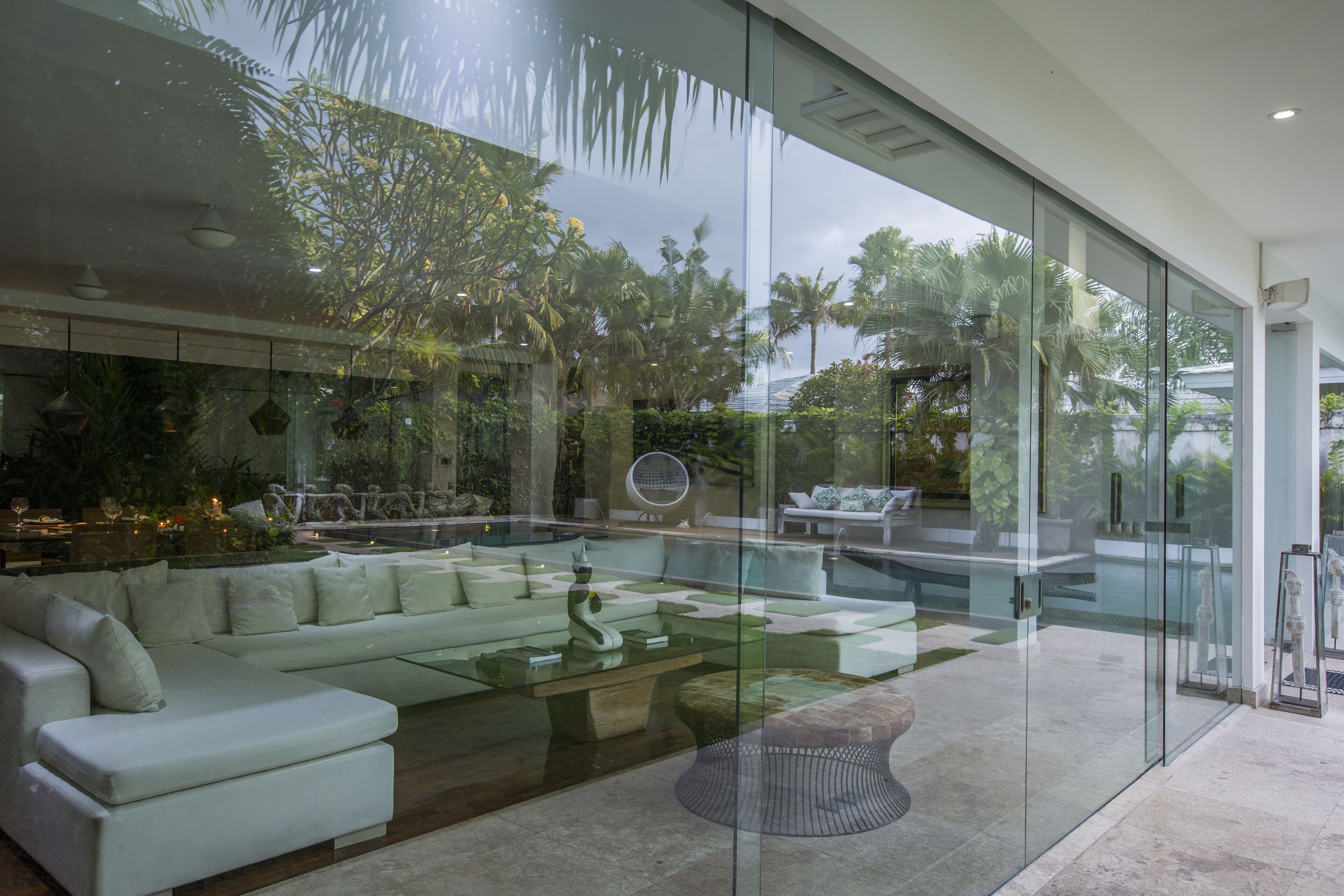 Una sala de estar moderna con un ventanal, un sofá blanco, una mesa de centro de vidrio y una zona de piscina visible a través del cristal.