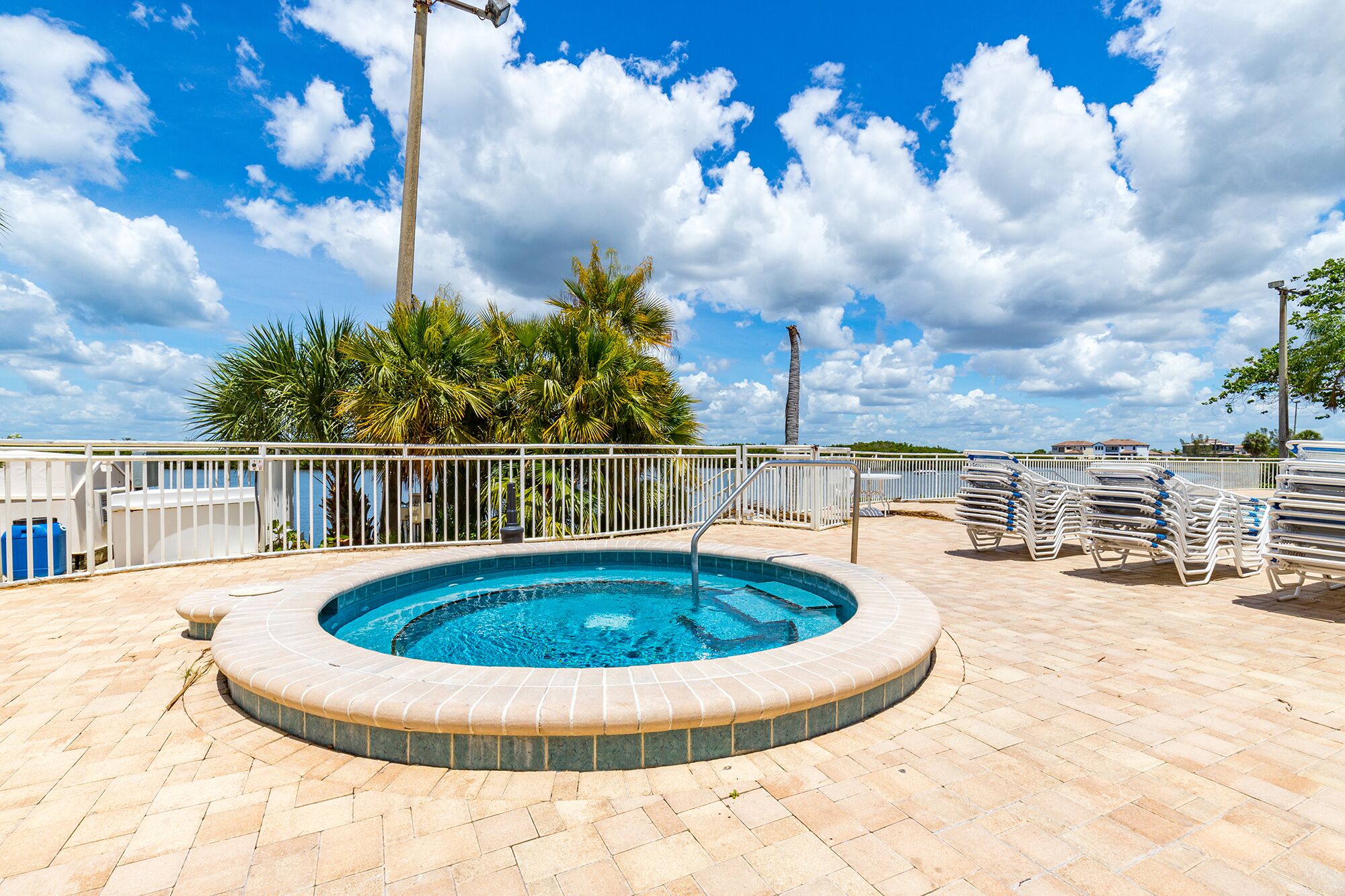 A circular pool with a brick border, surrounded by a white fence, palm trees, and a clear blue sky.