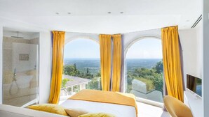 Ein modernes Schlafzimmer mit einem großen Bett, zwei Fenstern mit gelben Vorhängen, einem Fernseher und Blick auf die Landschaft.