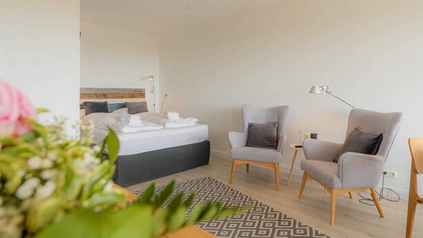 A modern bedroom with a bed, two armchairs, a side table, and a patterned rug. - Apartment 84 Coastal heart (ID 407) (Sankt Peter-Ording)