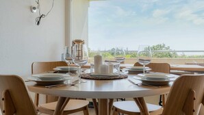A round dining table set with plates, glasses, and a candle centerpiece, surrounded by wooden chairs, with a view of greenery and a body of water through a large window. - Apartment 84 Coastal heart (ID 407) (Sankt Peter-Ording)