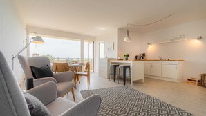 A modern living room with a grey sofa, a patterned rug, and a dining area with wooden chairs and a table. - Apartment 84 Coastal heart (ID 407) (Sankt Peter-Ording)