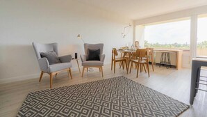 A spacious living area with two grey armchairs, a dining table with chairs, a patterned rug, and a large window offering a view of the outdoors. - Apartment 84 Coastal heart (ID 407) (Sankt Peter-Ording)