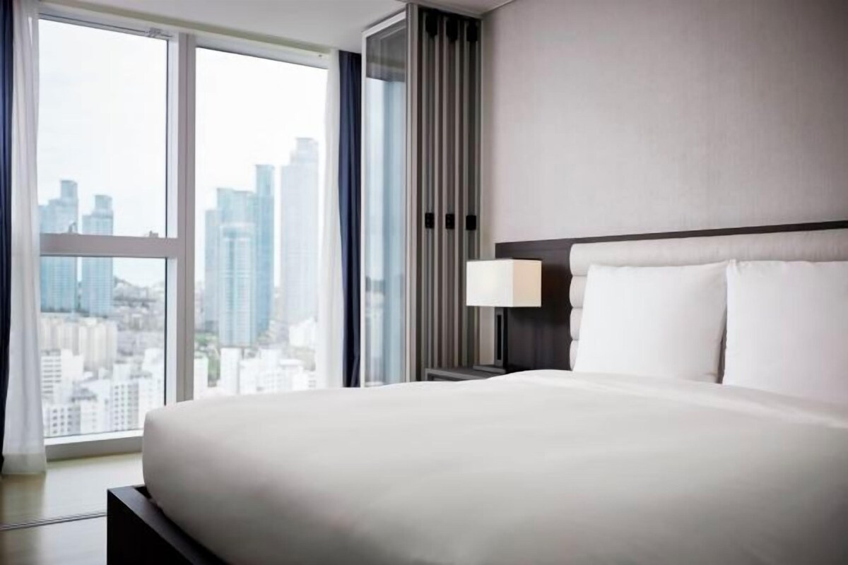 A hotel room with a large bed, a nightstand with a lamp, and a view of the cityscape through large windows.