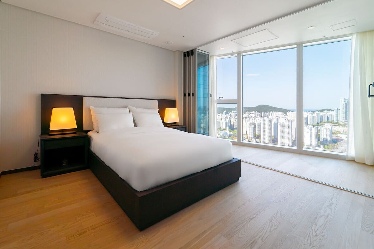 Un dormitorio moderno con una cama grande, dos mesitas de noche con lámparas y vistas a la ciudad a través de ventanales de gran altura.