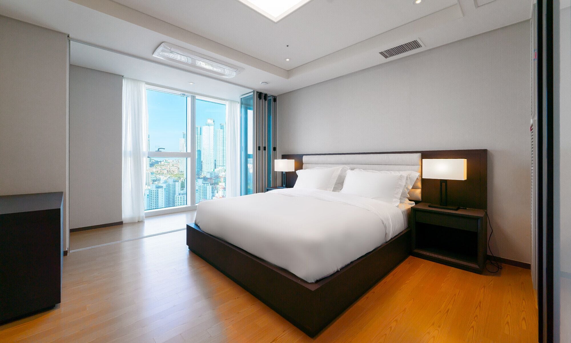 A modern hotel room with a large bed, two bedside tables with lamps, a view of the city through large windows, and a skylight.