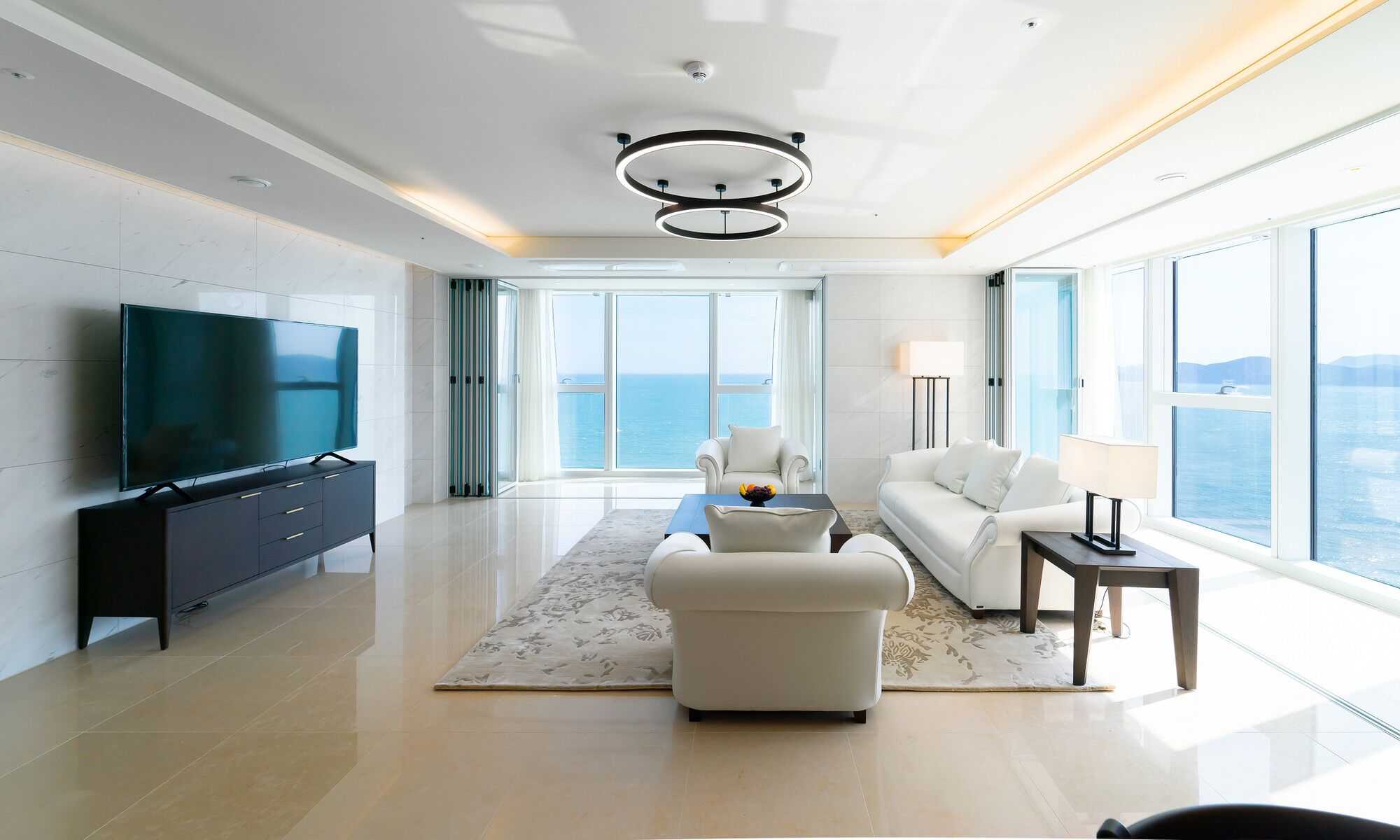 A modern living room with a large flat-screen TV, a sofa, a coffee table, and a view of the ocean through large windows.