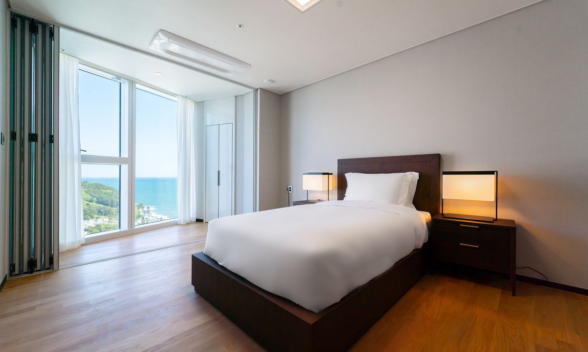 A modern hotel room with a large bed, two bedside lamps, a view of the ocean, and a sliding glass door.