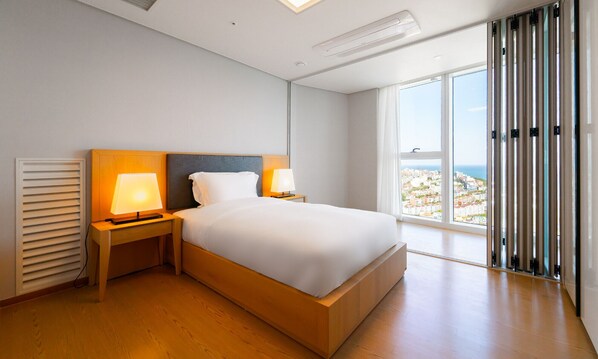 A modern hotel room with a large bed, two bedside lamps, a window with a city view, and a ceiling vent.