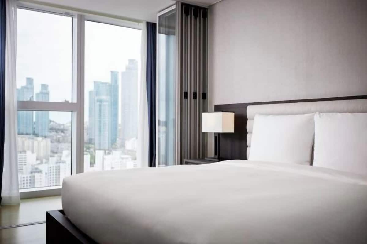 Habitación de hotel con una cama grande, una mesita de noche con lámpara y vistas a la ciudad a través de amplios ventanales.