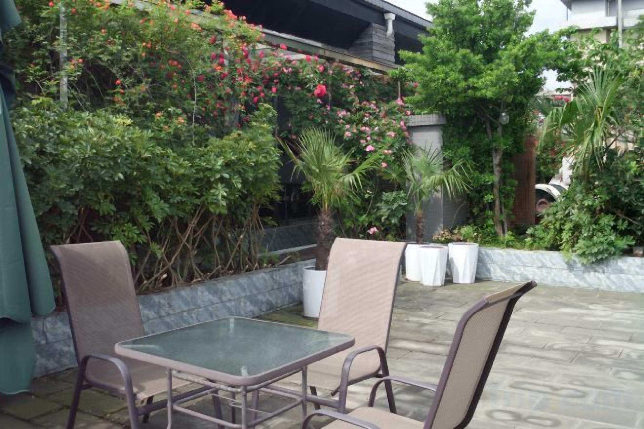 Un patio exterior con mesa y sillas, rodeado de abundante follaje y plantas con flores.